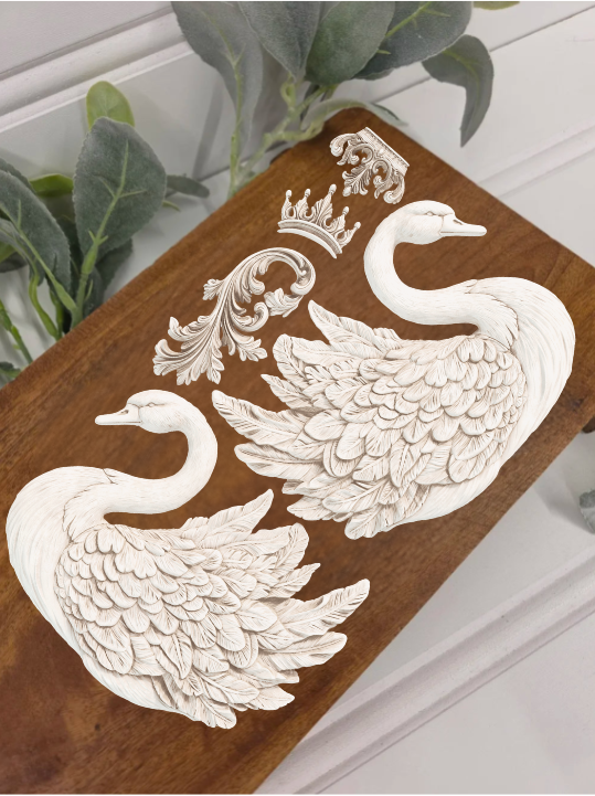 A rectangular wooden decorative board featuring two raised white swan figures and ornate scrollwork, photographed on a light surface with a small plant visible in the upper left corner.
