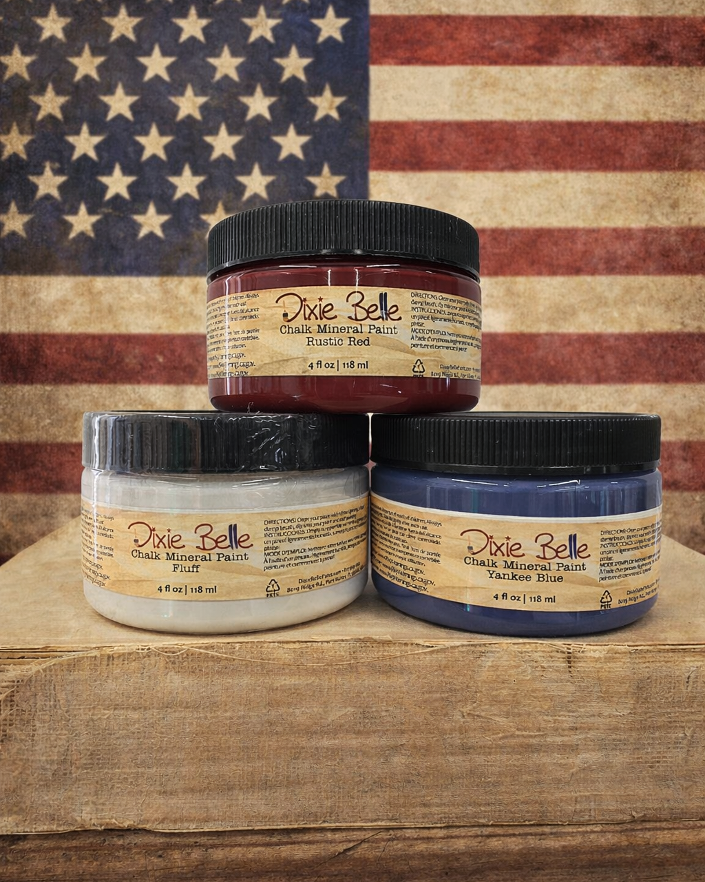 Three jars of Dixie Belle Chalk Mineral Paint stacked on a wooden surface with an American flag in the background.