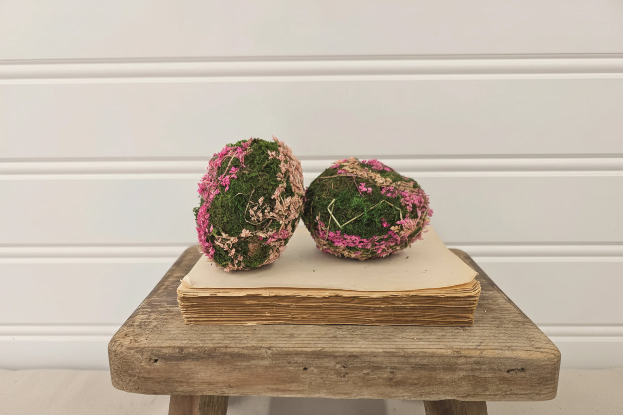 Two decorative moss balls with pink flowers on a wooden stool against a white wall.