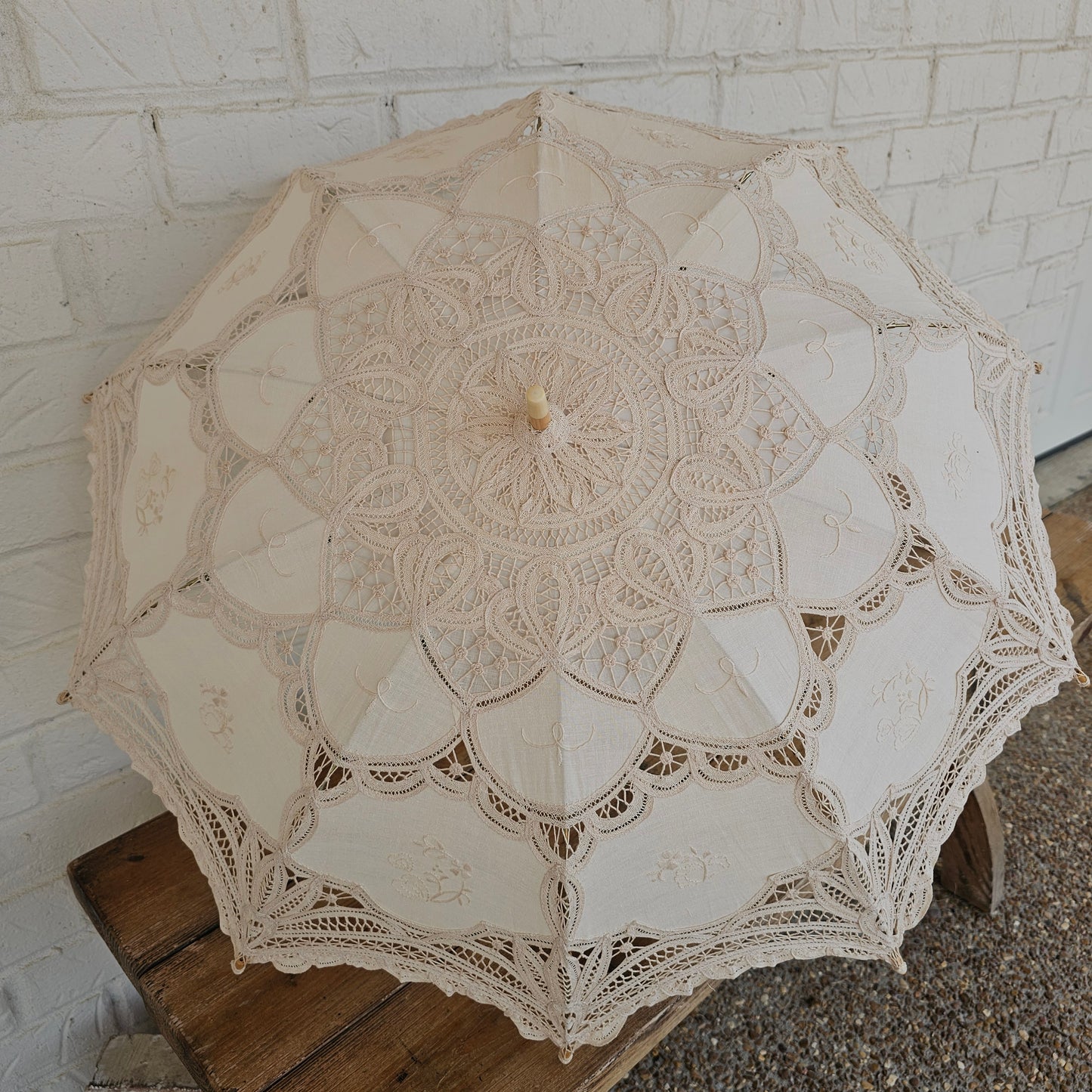 An off-white lace parasol umbrella with intricate floral and mandala patterns, shown from a top-down angle on a white brick patio next to a wooden bench.