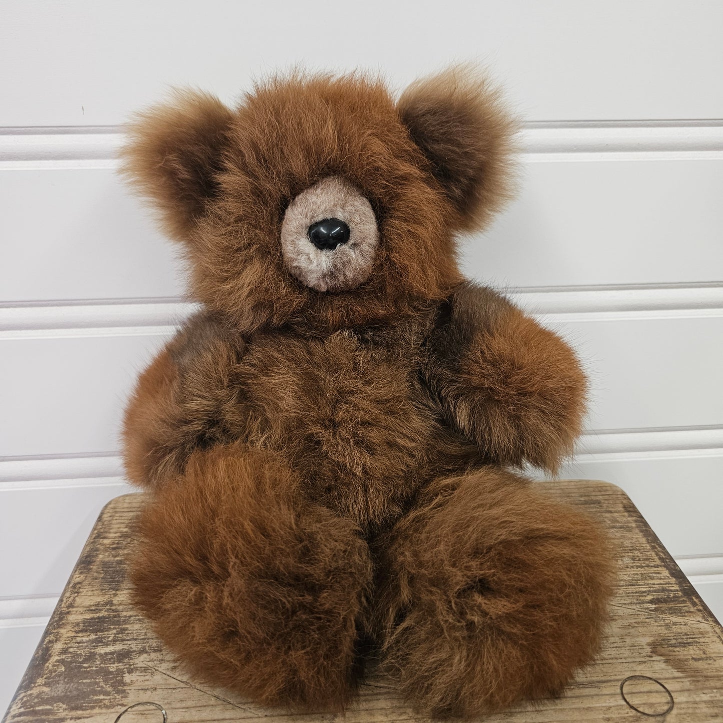 A fluffy medium brown teddy bear with a cream-colored snout and black nose, sitting on a rustic wooden stool against a white paneled wall.