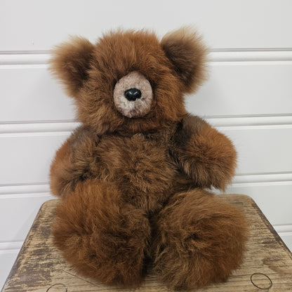 A fluffy medium brown teddy bear with a cream-colored snout and black nose, sitting on a rustic wooden stool against a white paneled wall.