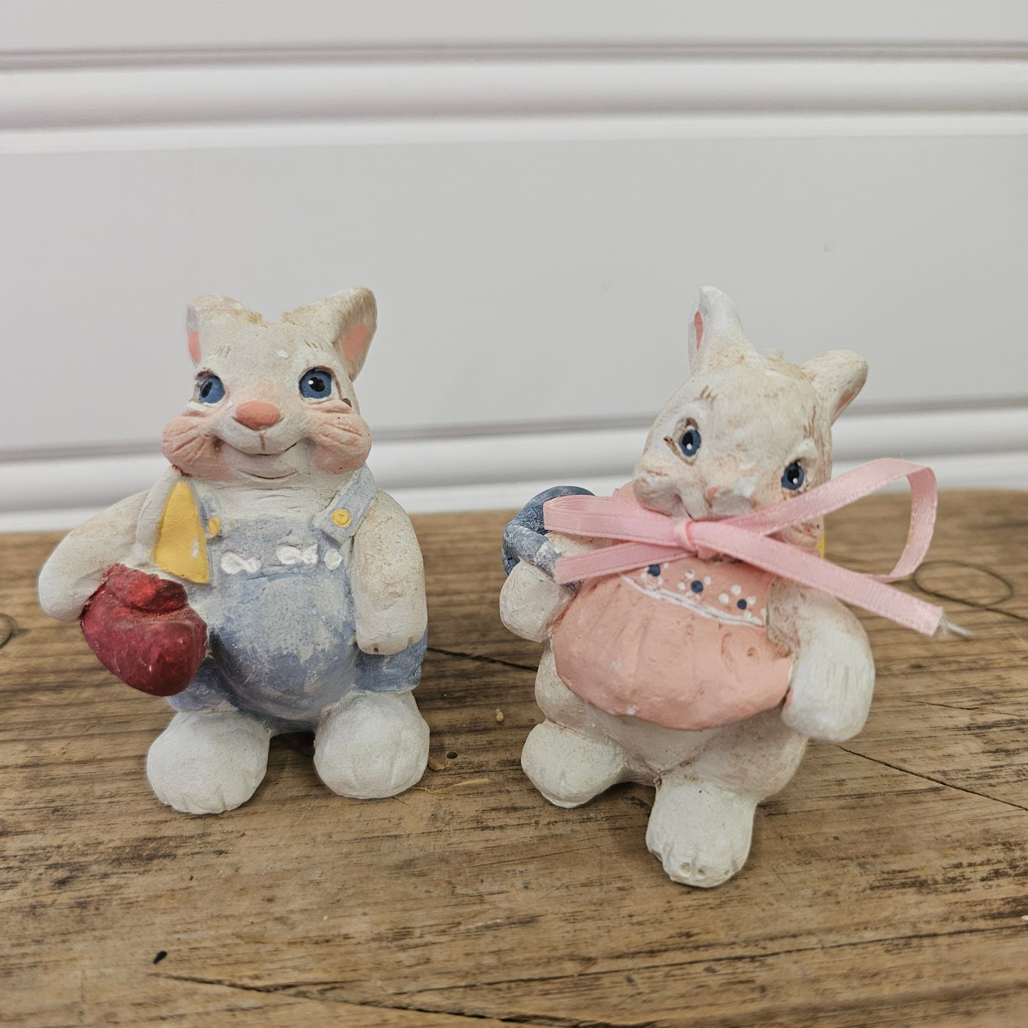 Two small decorative cat figurines posed on a wooden surface. One figurine is cream-colored with a pink bow and pink heart, and the other is cream-colored with blue overalls and a red heart.