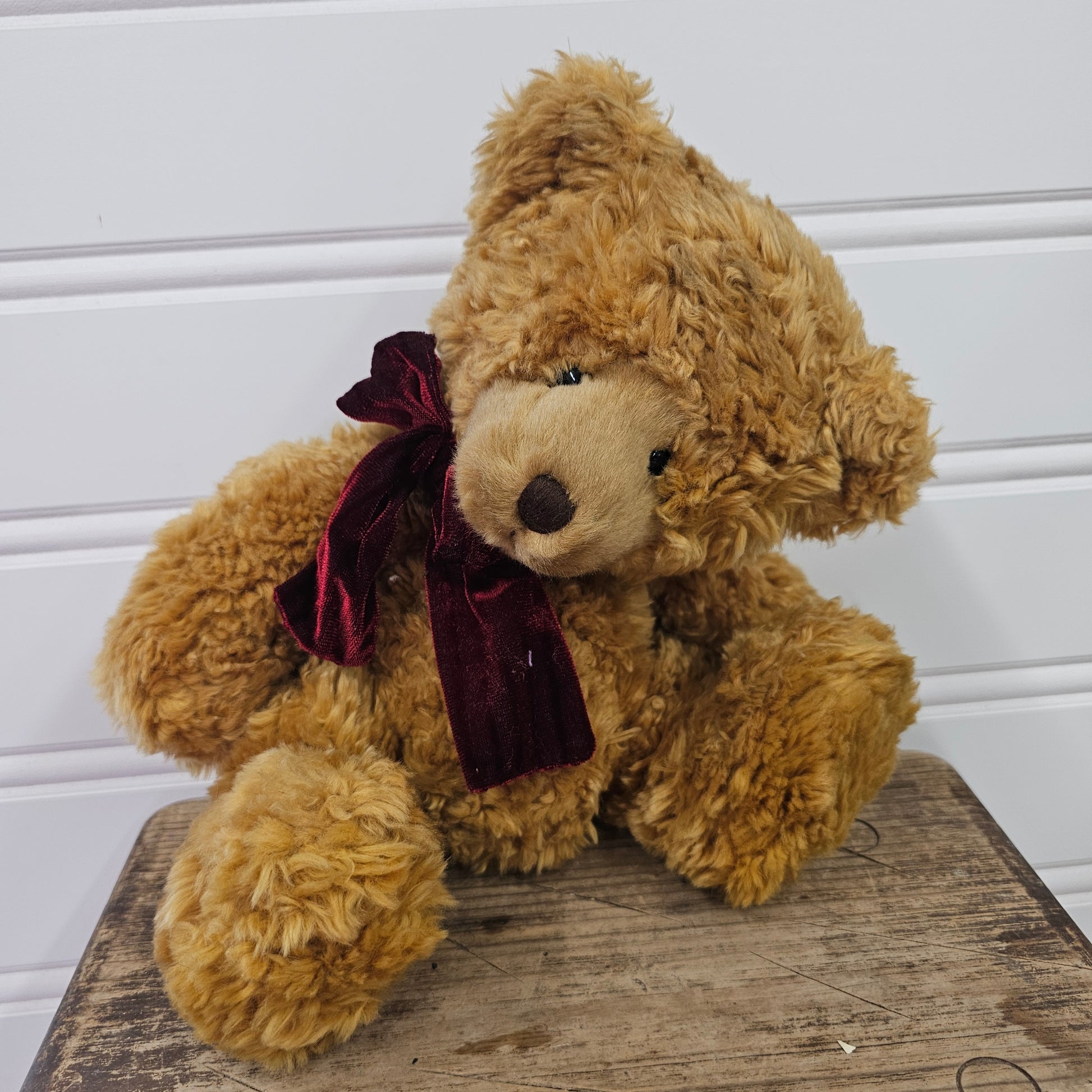 A plush teddy bear with curly golden-brown fur and a dark burgundy velvet bow around its neck, sitting on a rustic wooden surface against a white paneled wall.