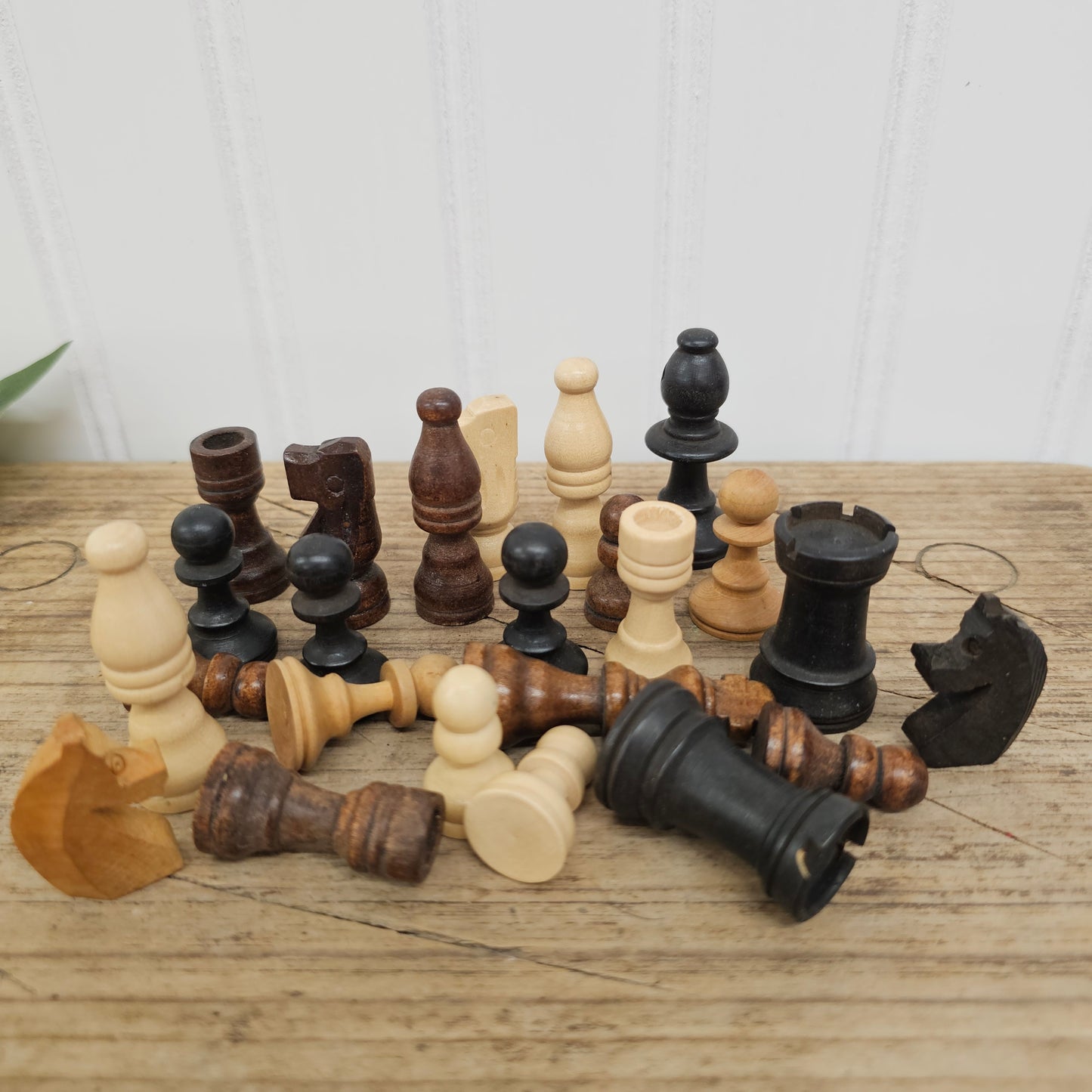 A set of vintage wooden chess pieces in natural light wood and dark black wood, arranged on a rustic wooden tabletop against a white paneled wall.