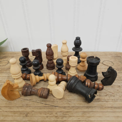 A set of vintage wooden chess pieces in natural light wood and dark black wood, arranged on a rustic wooden tabletop against a white paneled wall.