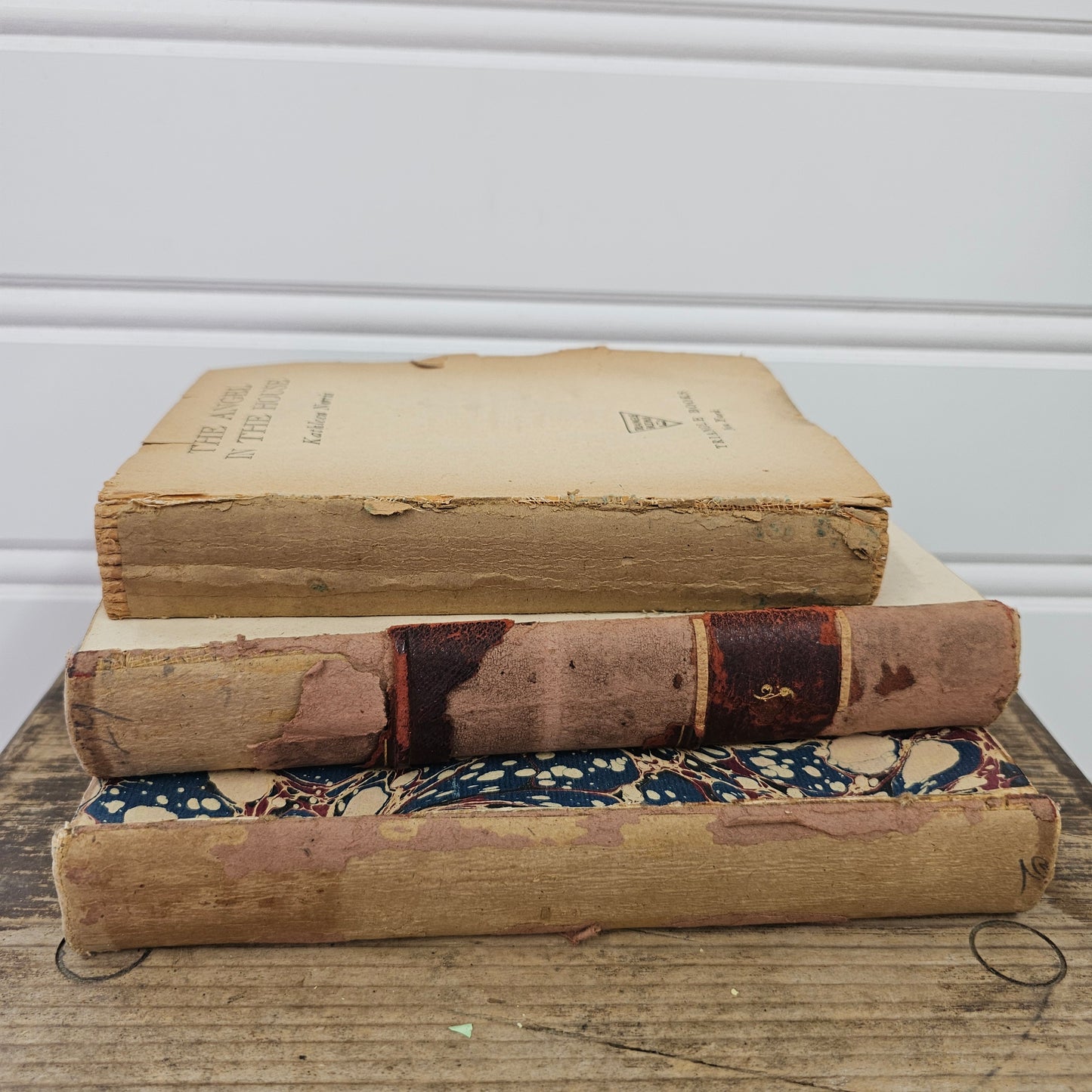 Three vintage, worn leather-bound books with faded tan and brown spines, displayed vertically on a rustic wooden surface against a white paneled wall.