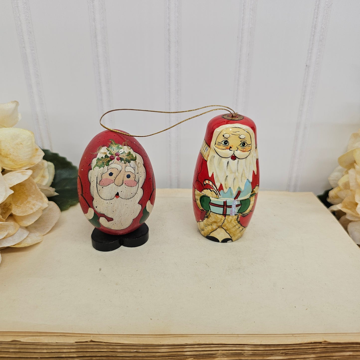Two hand-painted wooden Christmas ornaments shaped like Santa Claus, one standing and one egg-shaped, displayed on a vintage book page with white flowers in the background.