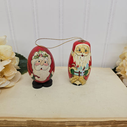 Two hand-painted wooden Christmas ornaments shaped like Santa Claus, one standing and one egg-shaped, displayed on a vintage book page with white flowers in the background.